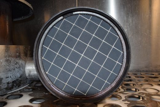 Filtre à air rond noir avec une grille grise, posé sur une surface métallique et photographié en gros plan.