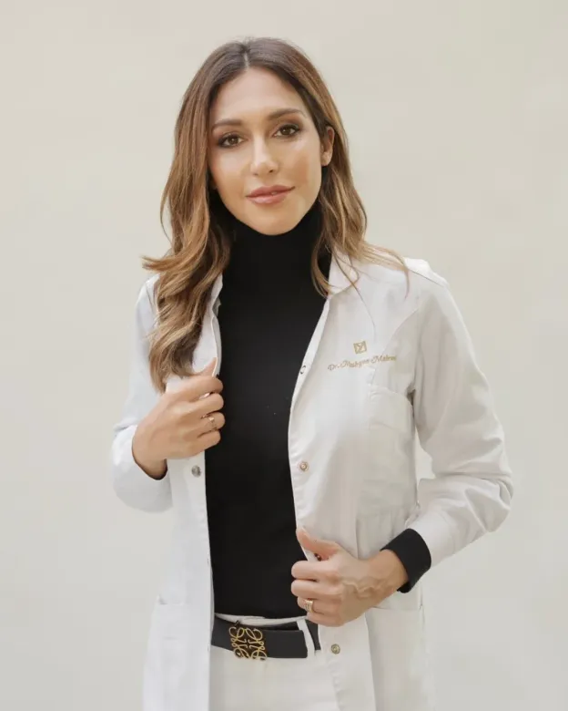 Una mujer lleva una bata de laboratorio blanca y un jersey de cuello alto negro.