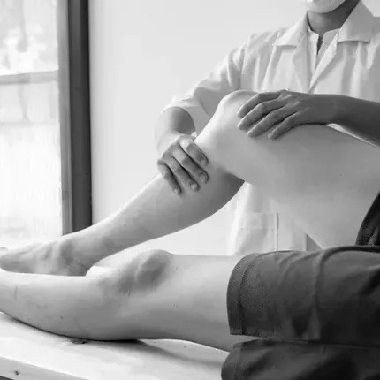 Un profesional médico manipula la rodilla de un paciente en el consultorio. El paciente yace en una camilla mientras el médico viste una bata.