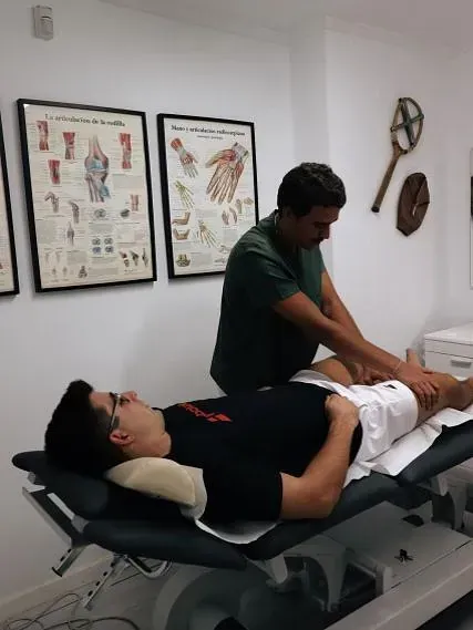 Un hombre con camisa verde masajea la pierna de un hombre en una mesa de operaciones en una clínica. Hay tablas de anatomía colgadas en la pared.