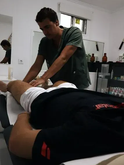 Un hombre con uniforme verde masajea la pierna de una persona tumbada en una camilla. El ambiente parece el de una clínica o un spa.