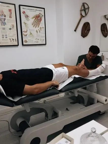 Un hombre yace boca abajo en una camilla mientras otro terapeuta le trabaja el cuello. La sala tiene historiales médicos en la pared.