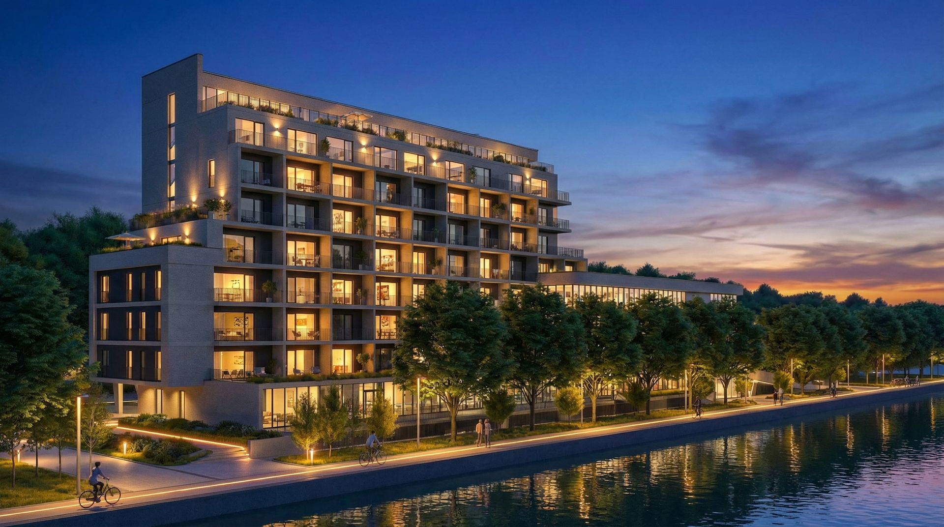 Immeuble moderne à plusieurs étages au crépuscule, fenêtres éclairées, le long d'un cours d'eau bordé d'arbres.