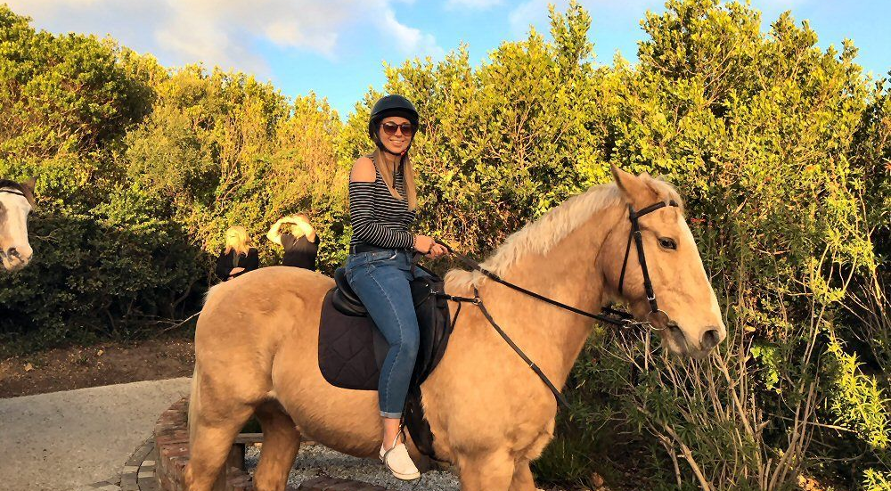 Woman in riding helmet on a tan horse outdoors, near greenery.