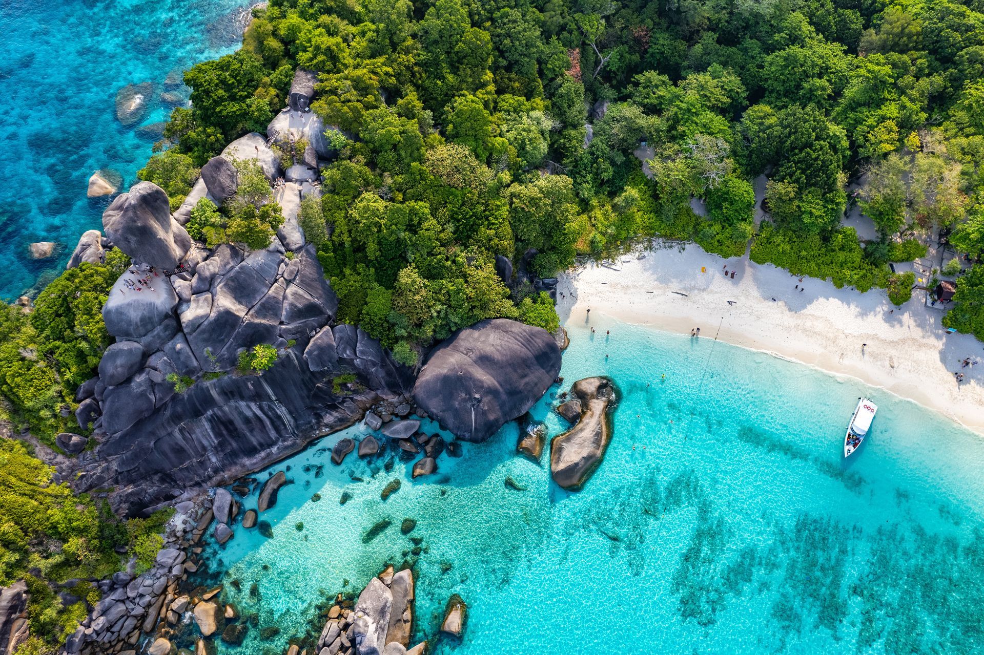 Aerial view of a tropical beach with turquoise water, white sand, lush green trees, and large gray rock formations.