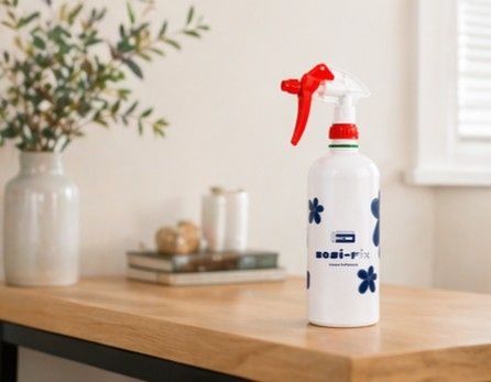 Un frasco pulverizador blanco con gatillo rojo sobre una mesa de madera en una habitación luminosa.