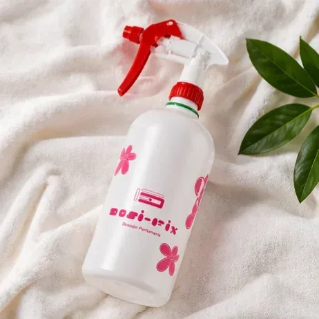 Botella pulverizadora blanca con boquilla roja y etiqueta floral rosa sobre un fondo de tela clara, junto a hojas verdes.