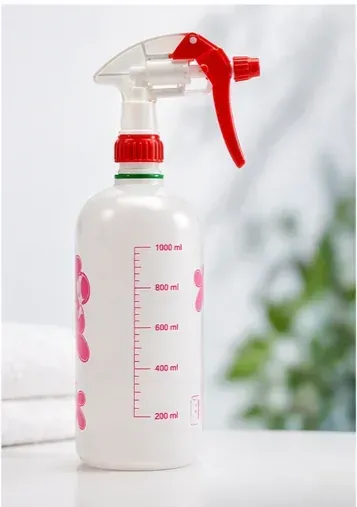 Botella pulverizadora transparente con gatillo rojo y marcas de medición sobre una superficie blanca.