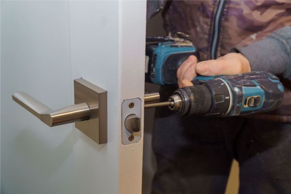Persona que utiliza un taladro para instalar una cerradura en una puerta blanca; herrajes plateados visibles.