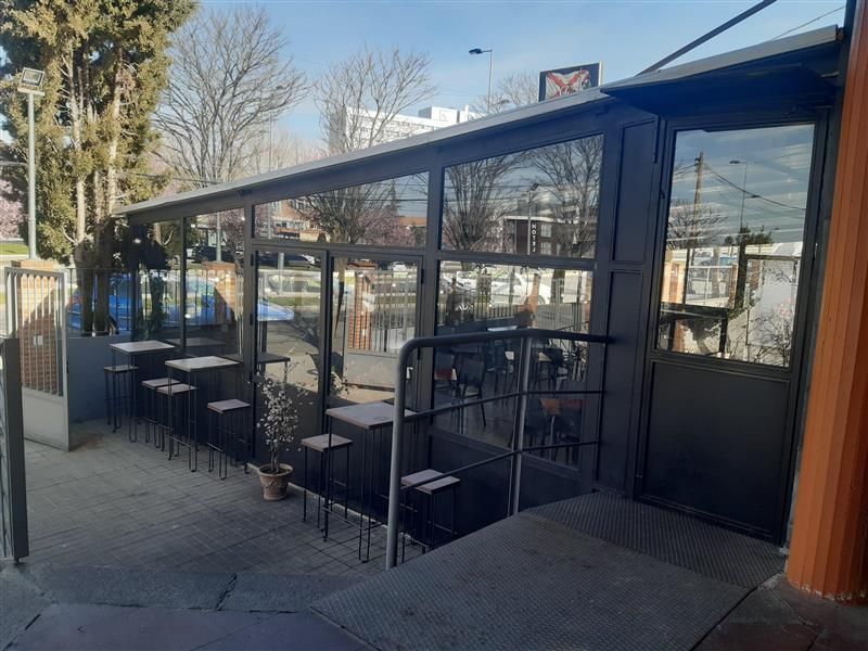 Un patio con una cafetería acristalada, mesas altas y taburetes, con el paisaje urbano diurno de fondo.