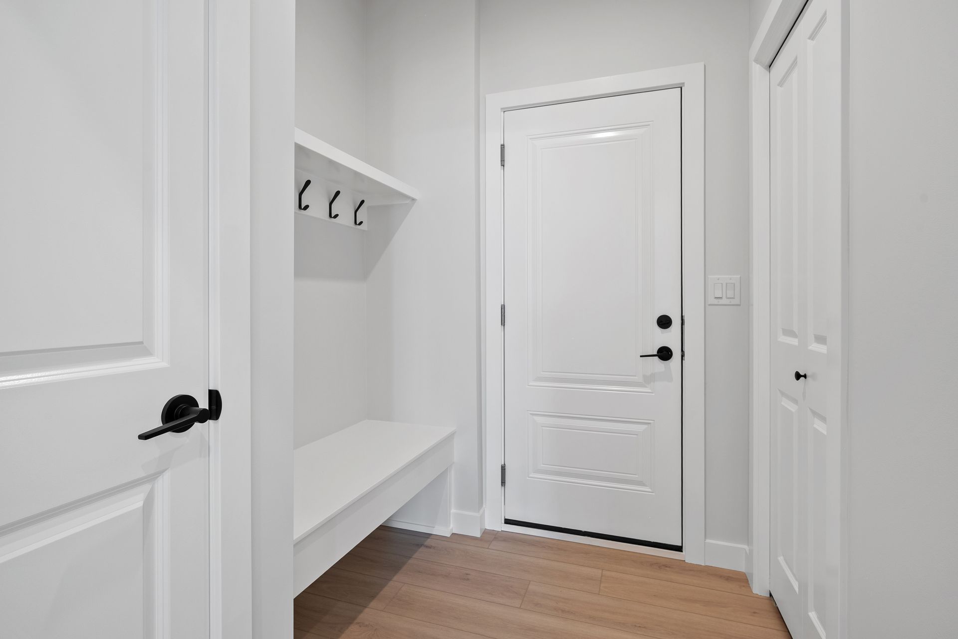 Un couloir blanc avec une banquette intégrée, des crochets et des portes ; parquet clair.