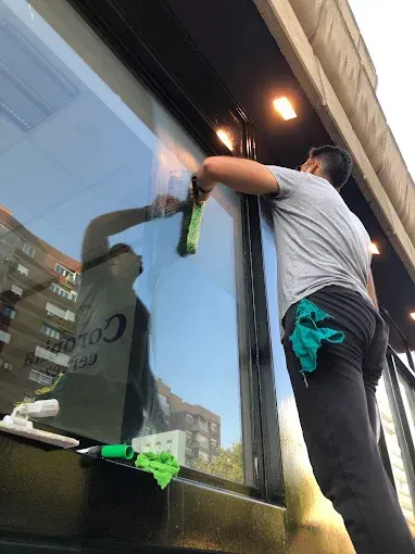 Una persona con camisa gris y pantalones oscuros limpia el escaparate de una tienda con un paño verde.