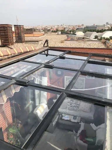 Un techo acristalado con vistas a una habitación amueblada, con el telón de fondo de una azotea urbana.