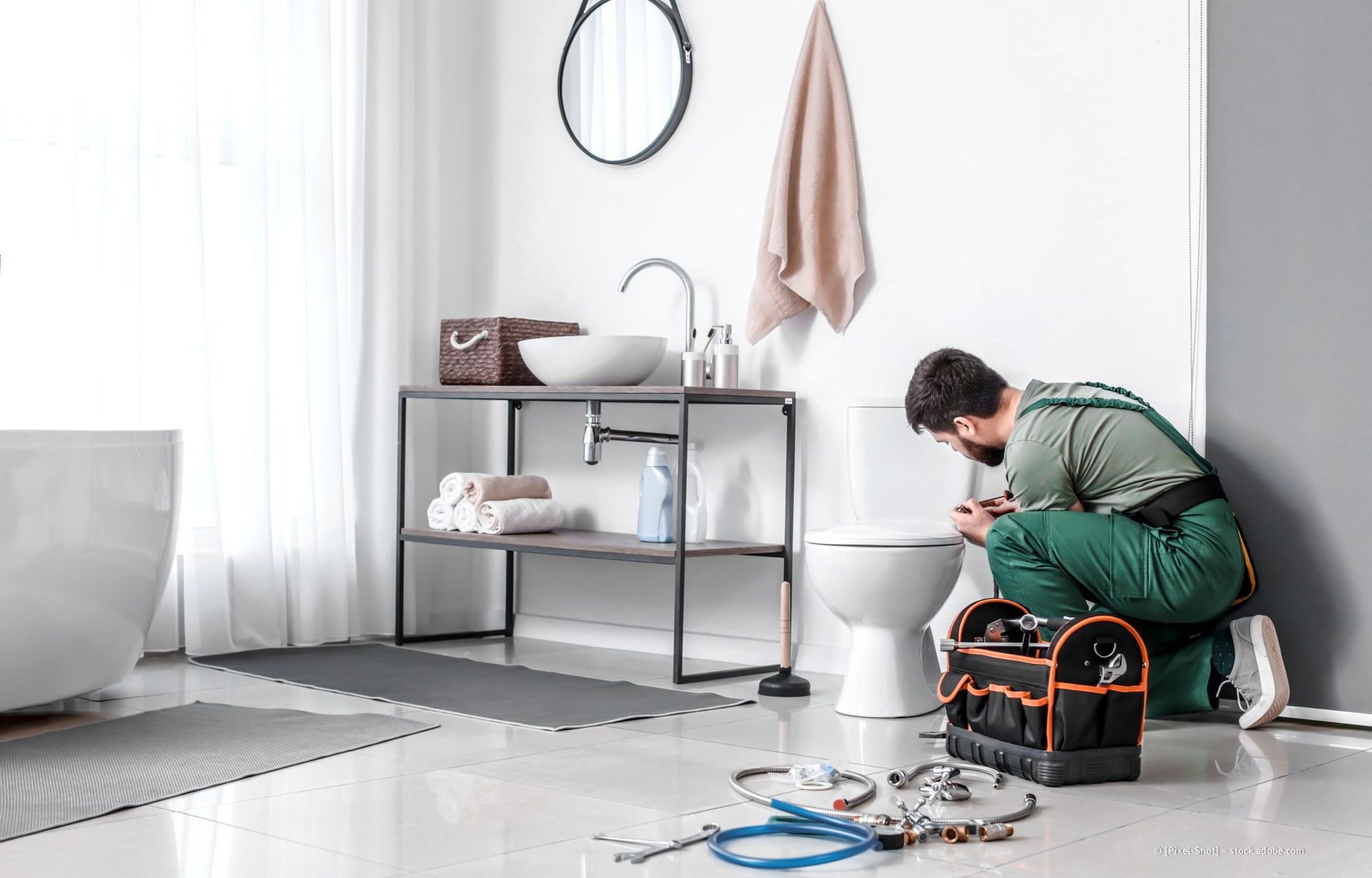 Ein Klempner repariert eine Toilette in einem modernen Badezimmer. Werkzeug und Zubehör liegen auf dem Boden.