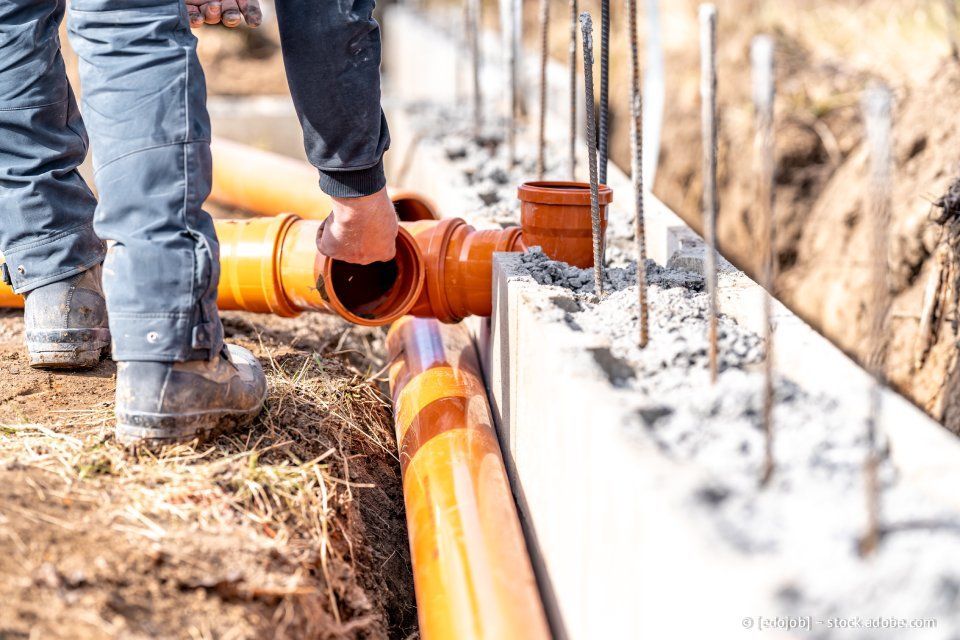 Ein Bauarbeiter verbindet orangefarbene Abflussrohre in der Nähe eines Betonfundaments.