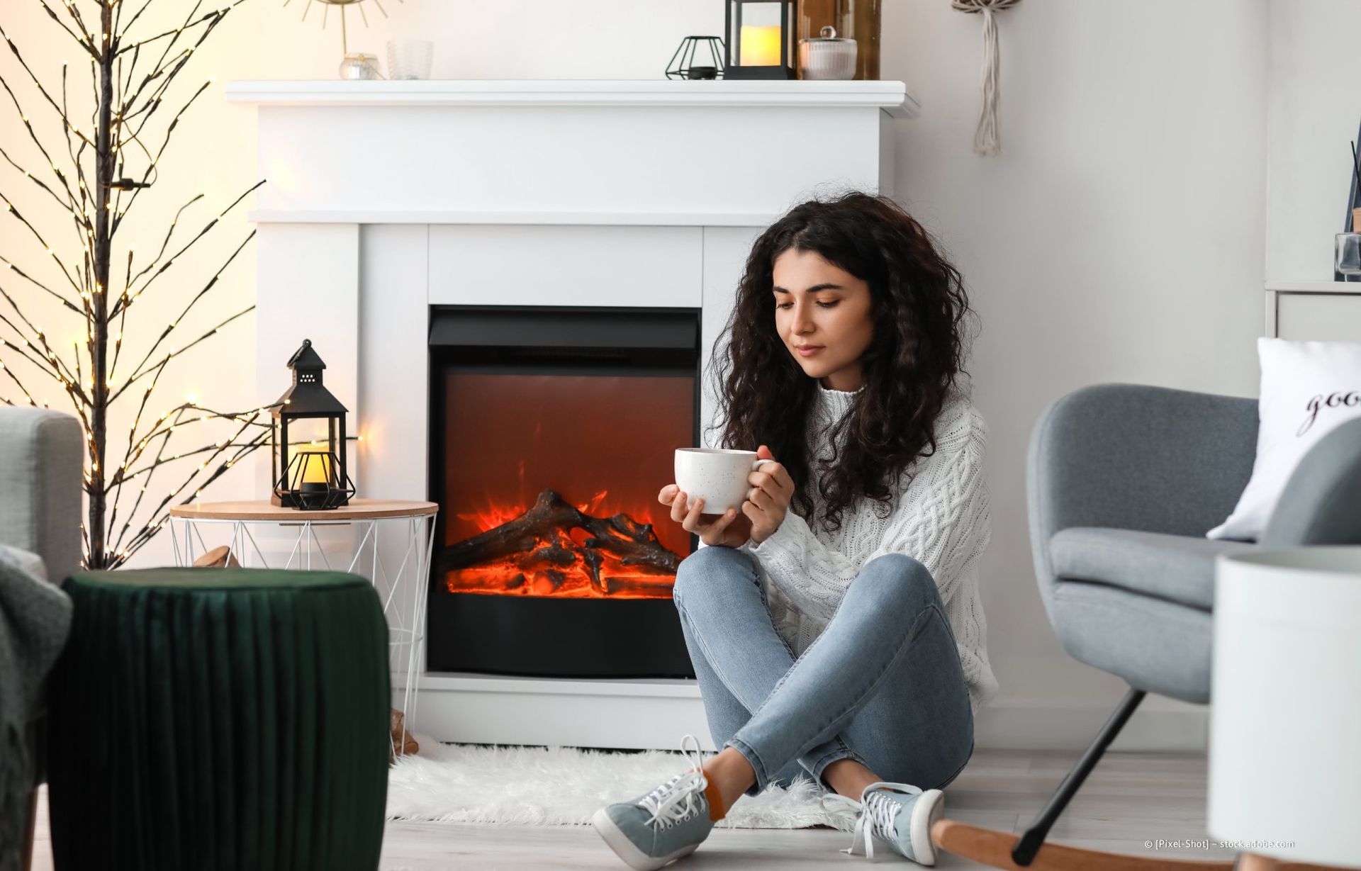 Eine Frau sitzt am Kamin und hält eine Tasse. Sie trägt Jeans und einen Pullover und wirkt nachdenklich. Das Wohnzimmer ist dekoriert.