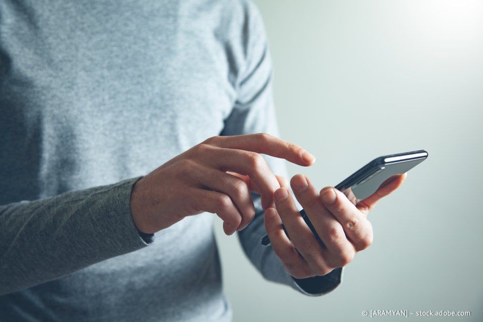 Eine Person, die ein Smartphone benutzt und mit den Fingern auf den Bildschirm tippt. Sie trägt ein graues Langarmhemd.