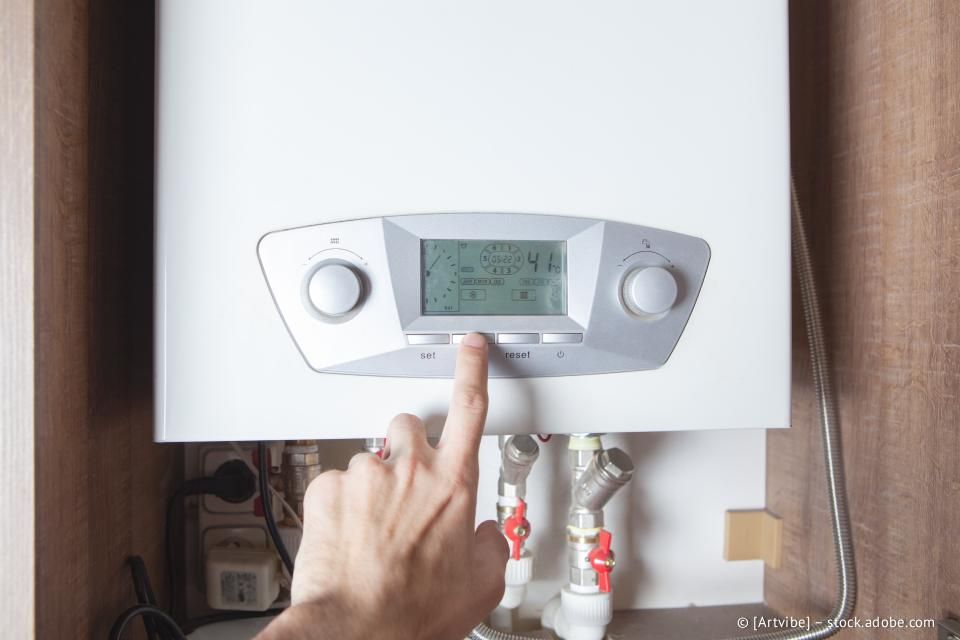 Eine Hand drückt einen Knopf auf einem weißen Heizkessel-Bedienfeld, das an einer Holzwand montiert ist.