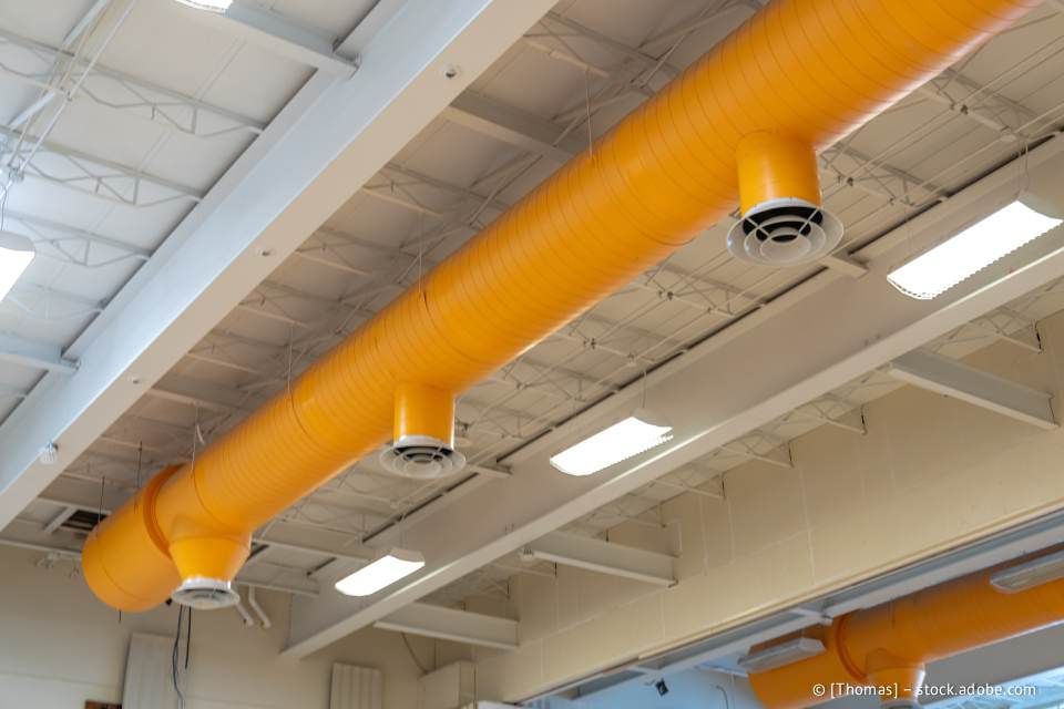 Orangefarbene, flexible Lüftungskanäle verlaufen über eine weiße Decke mit Lüftungsschlitzen.