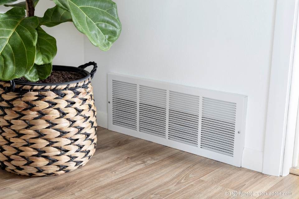 Neben einer weißen Wand mit einem rechteckigen Lüftungsschlitz auf einem Holzboden steht eine Topfpflanze.