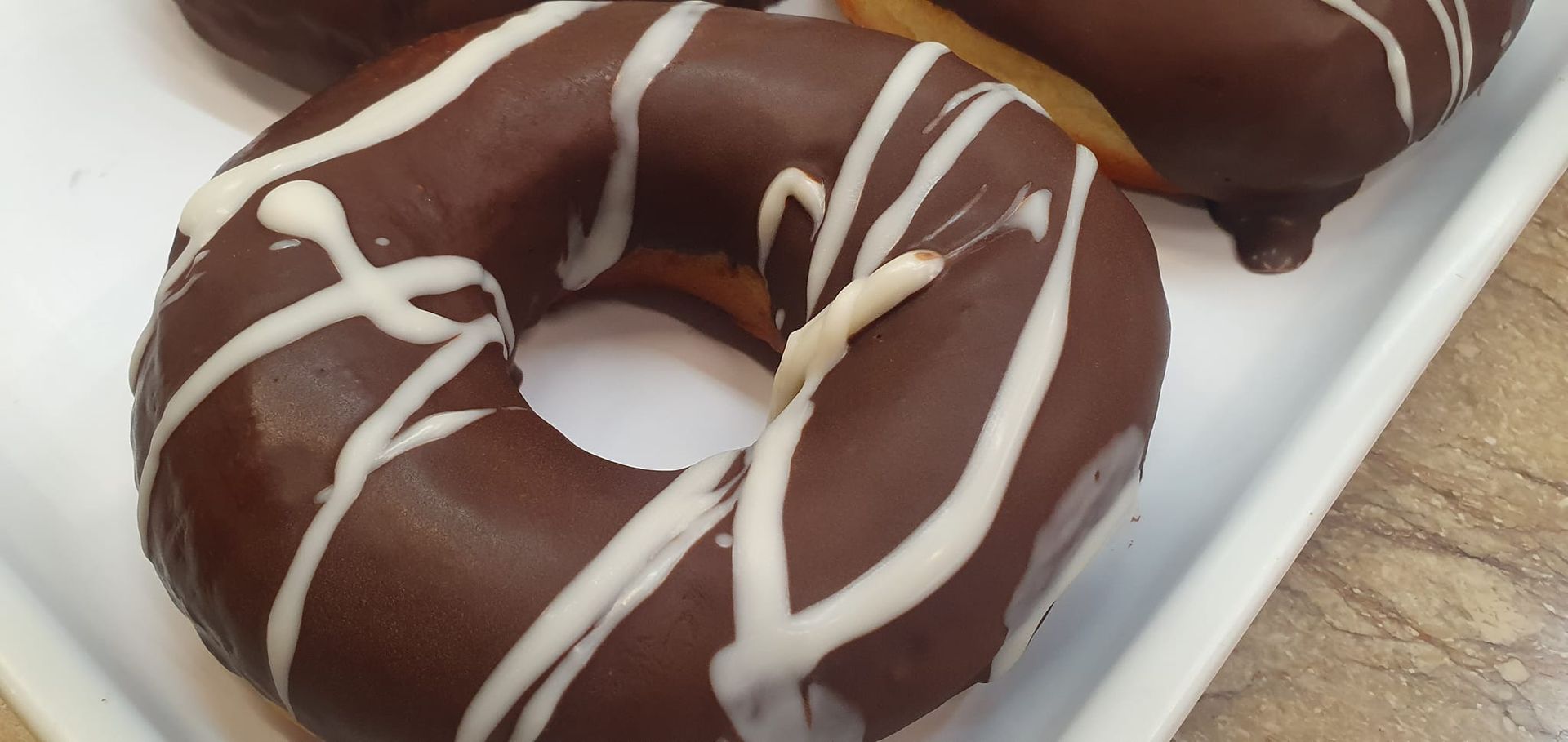 Primer plano de una rosquilla cubierta de chocolate con un chorrito de glaseado blanco sobre una bandeja blanca.