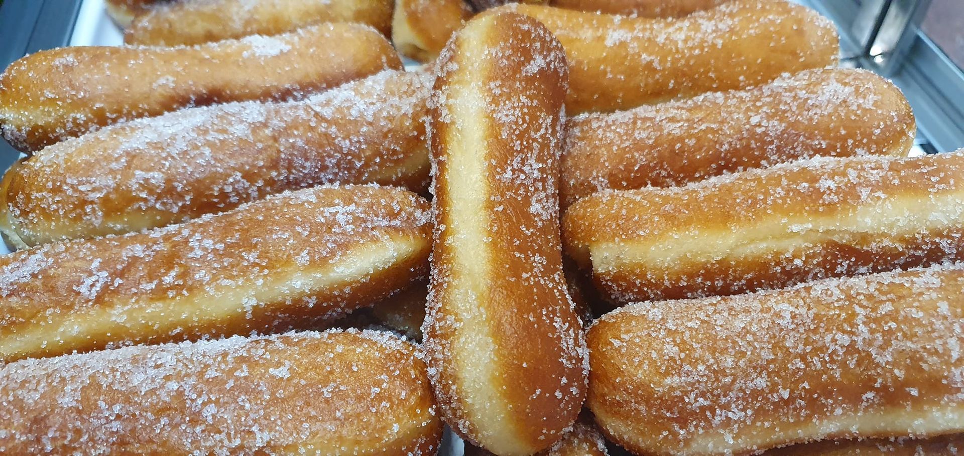 Primer plano de una bandeja llena de rosquillas fritas alargadas, de color marrón dorado, recubiertas de azúcar granulada.
