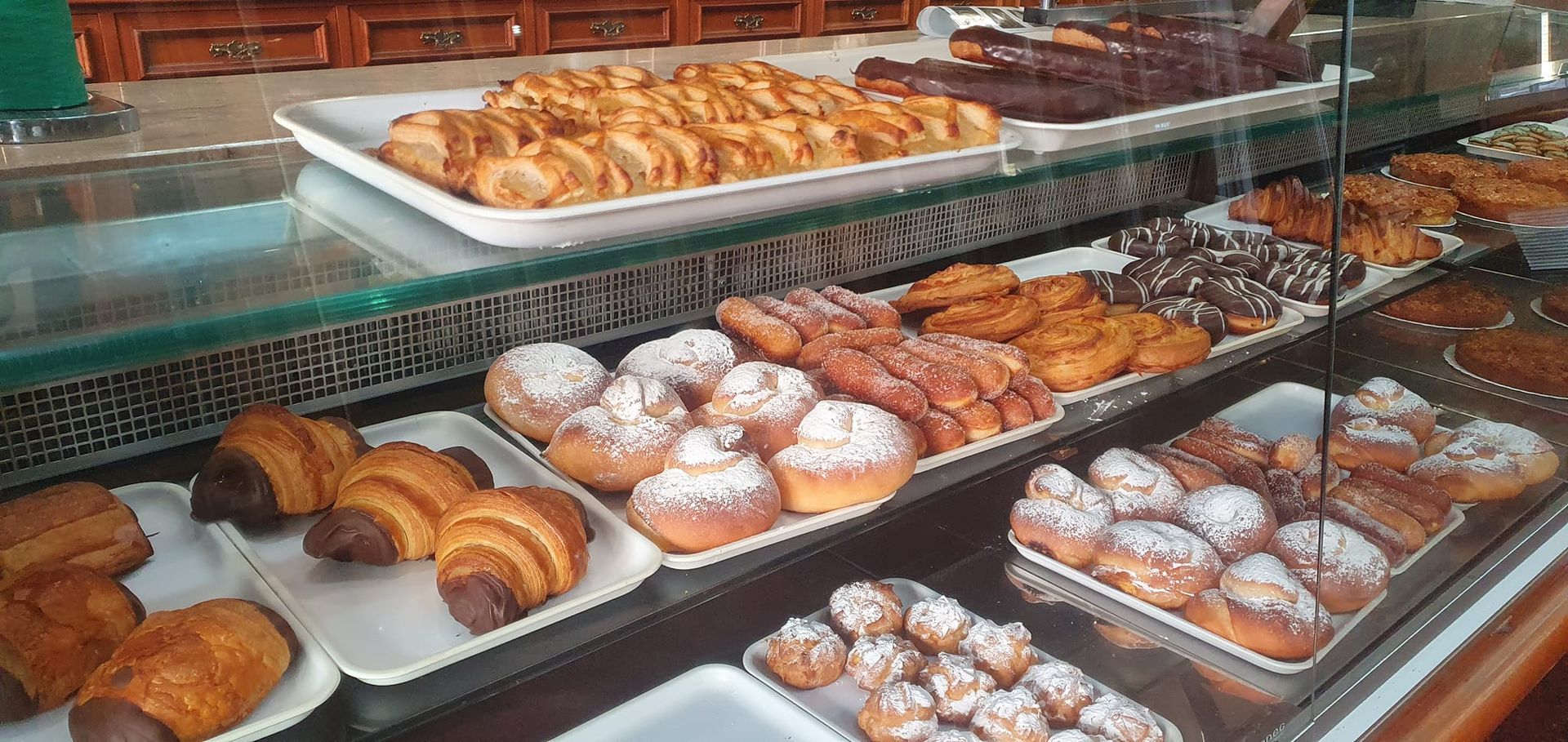Una vitrina en una panadería llena de diversas bandejas de pasteles recién hechos, incluyendo croissants, donas y panecillos.