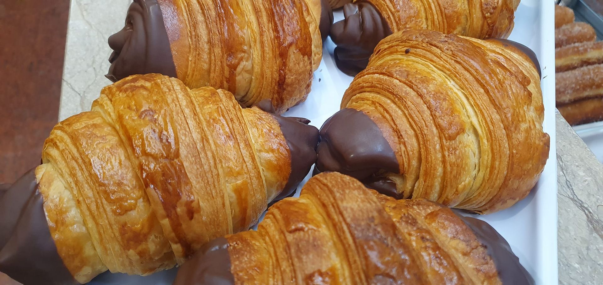Primer plano de varios croissants dorados y hojaldrados bañados en chocolate, dispuestos en una bandeja blanca.