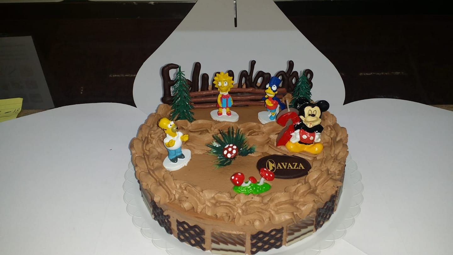 Una tarta de cumpleaños de chocolate decorada con figuritas de Los Simpson y Mickey Mouse, una vela roja y adornos festivos.
