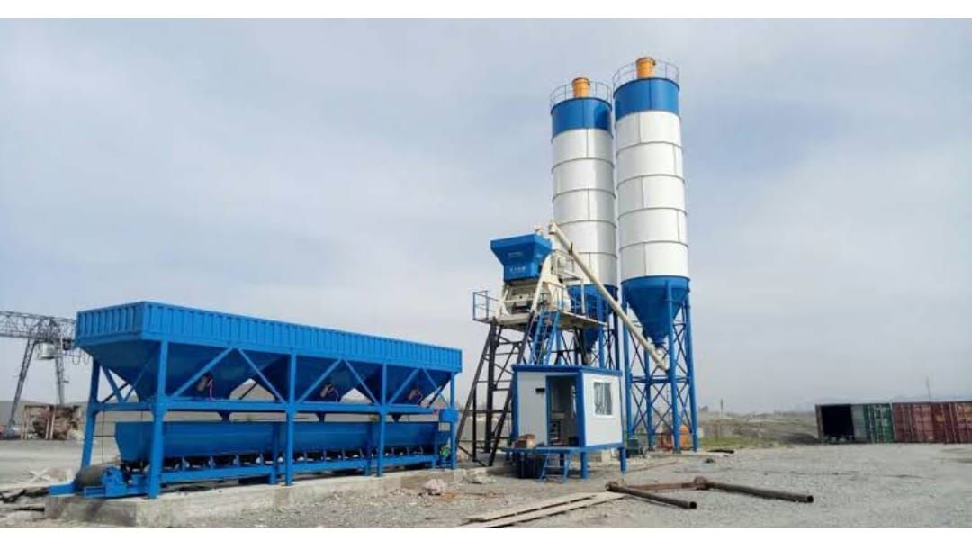 A concrete mixer is sitting in the middle of a dirt field next to two silos.