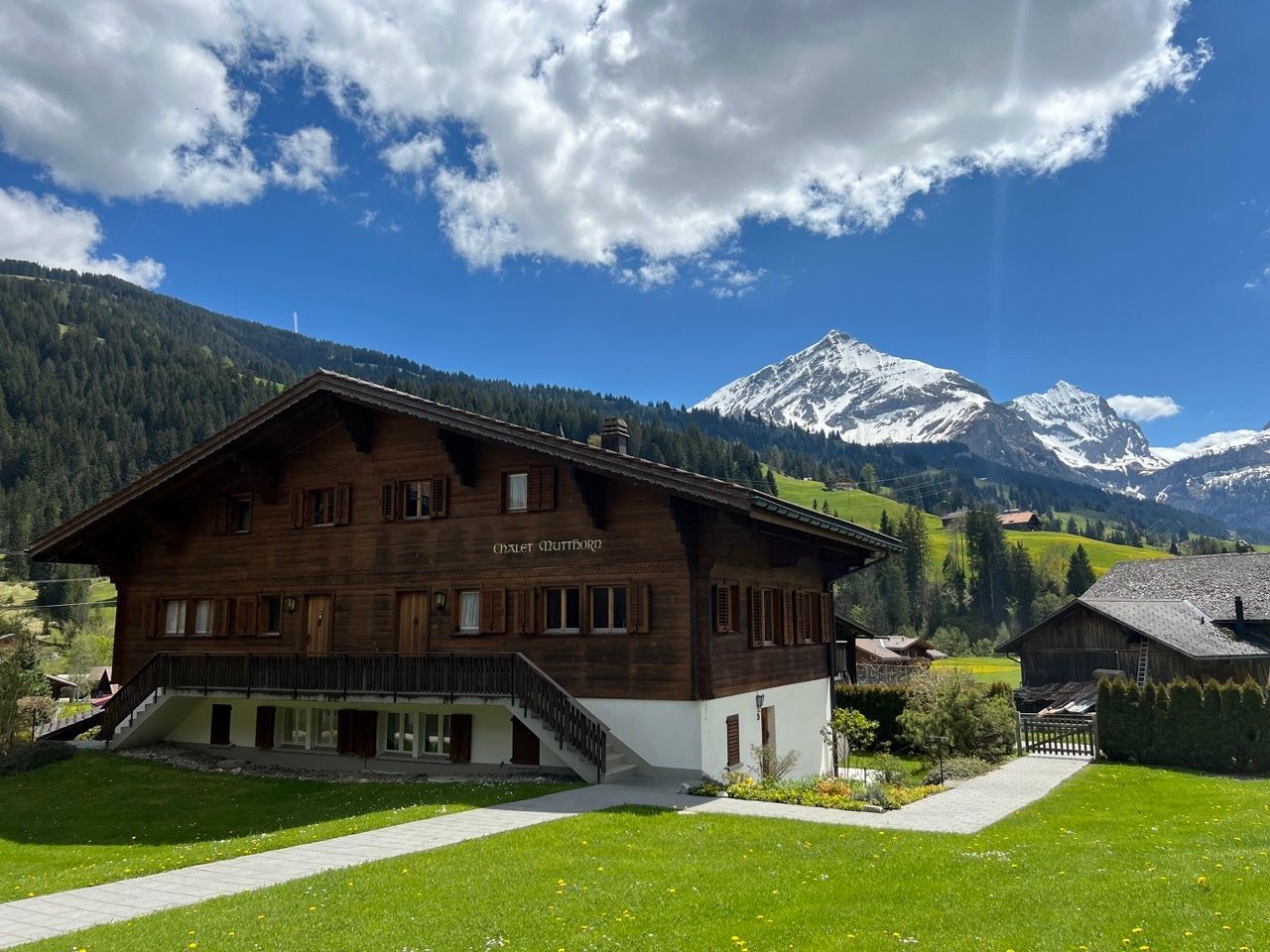 Wooden chalet in a mountain valley with green lawns, snowy peak, and blue sky with clouds