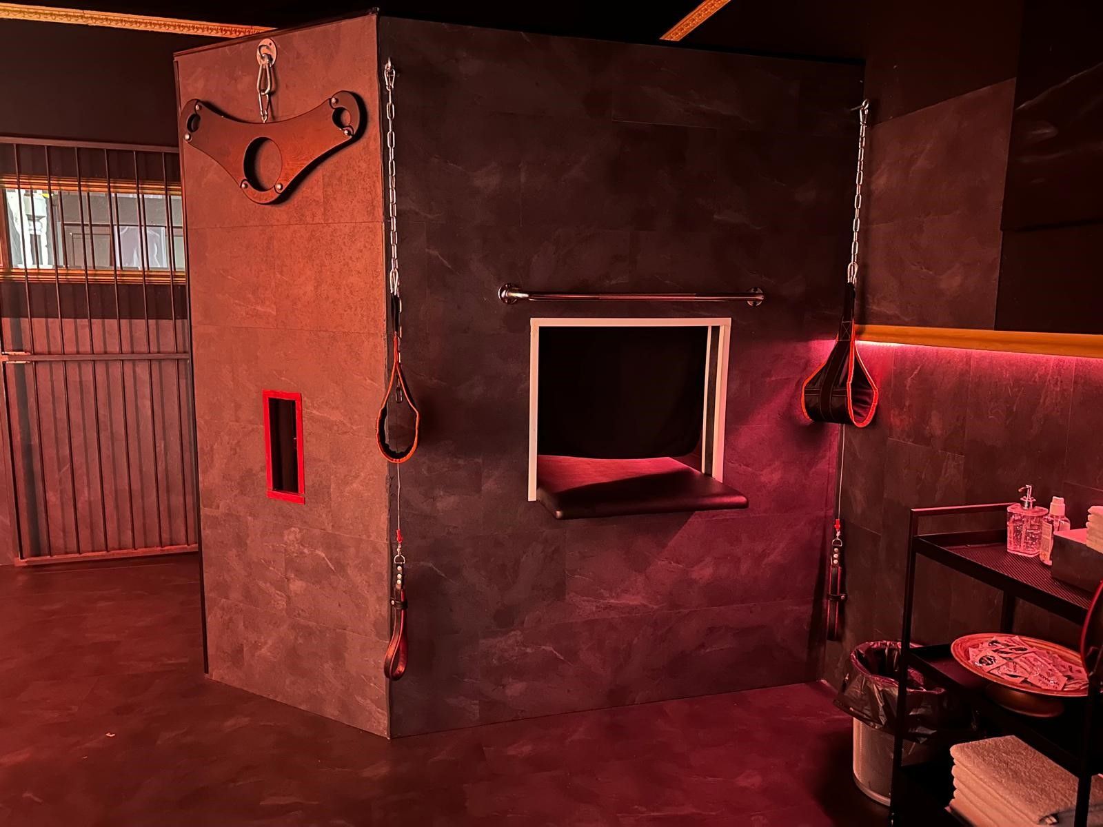 Dunkler Raum mit grauen Wänden, roter Beleuchtung, hängenden Fesseln, kleinem Fenster und schwarzen Metallregalen.