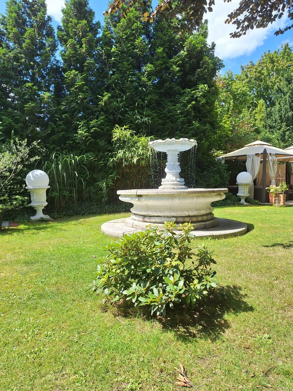 Üppig grüner Garten mit weißem Brunnen, dekorativen Urnen und einem kleinen Pavillon.