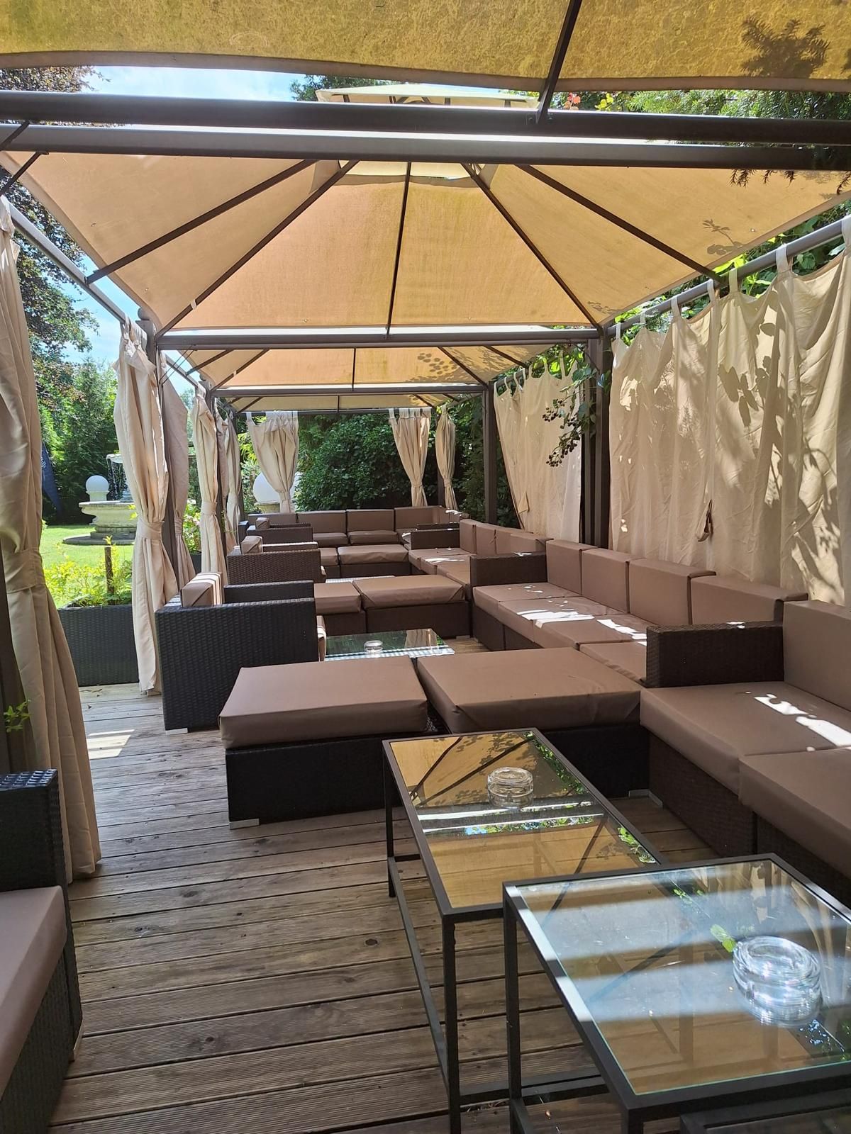 Lounge-Bereich im Freien unter einem Baldachin mit braunen Sofas, Glastischen und transparenten Vorhängen.