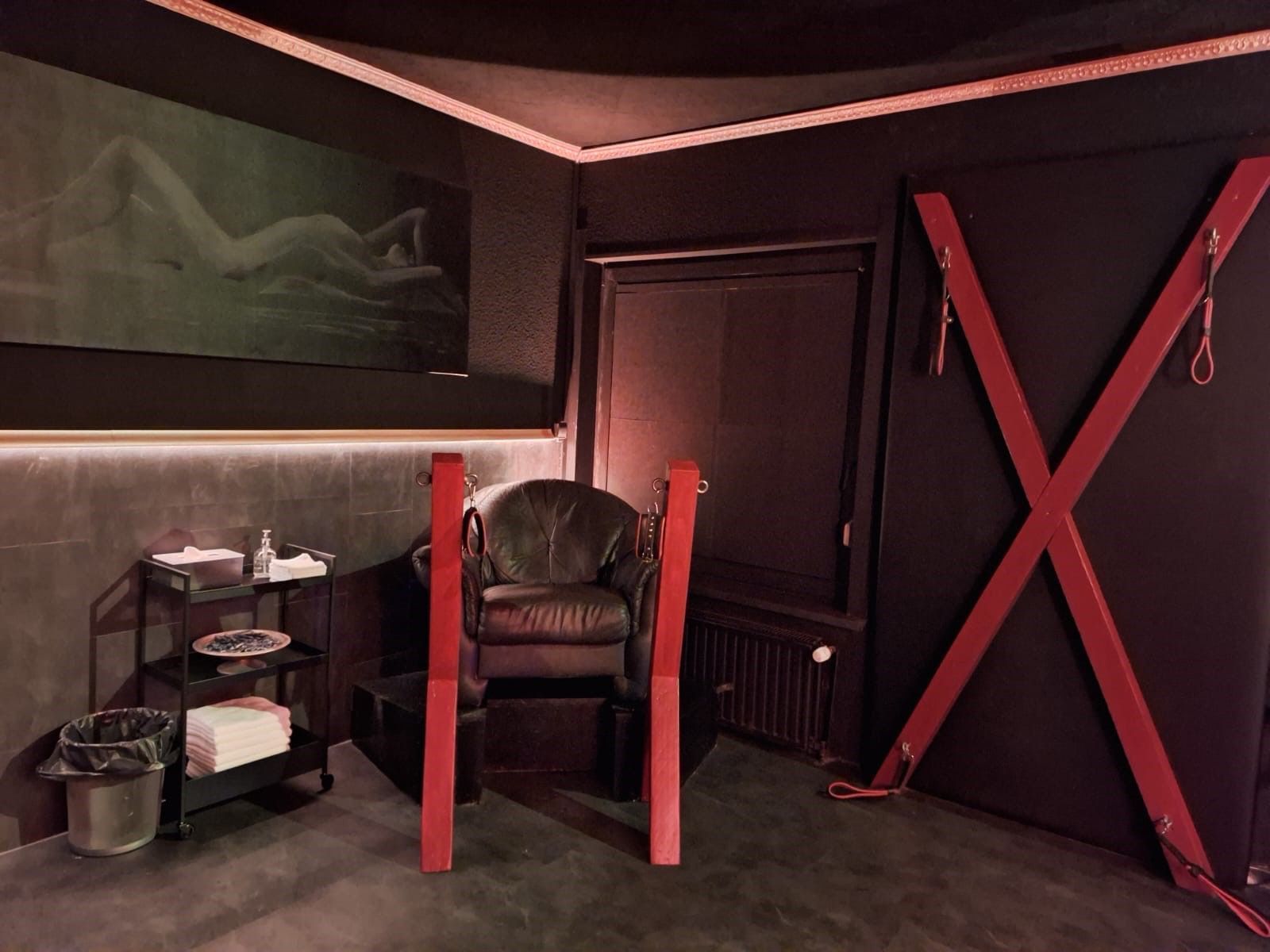Dunkler Raum mit roten Akzenten; ein Ledersessel, ein Kreuz und ein Gemälde sind zu sehen.