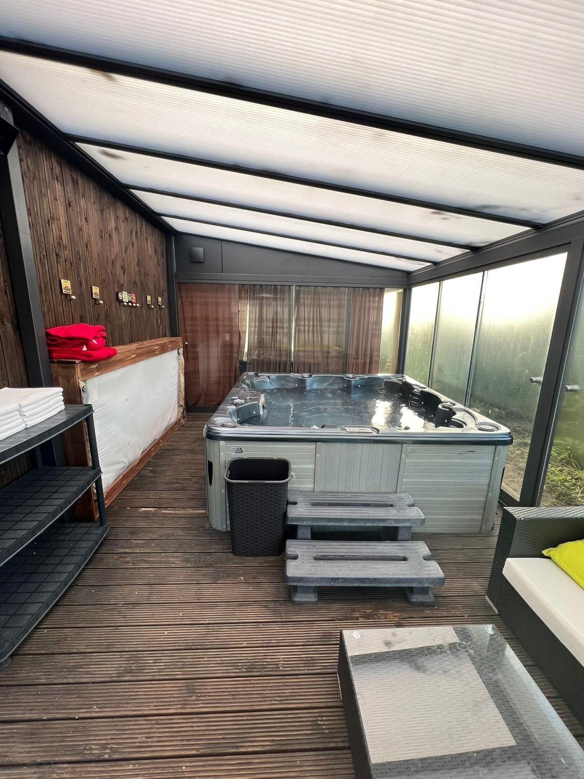 Swim-Spa unter einer überdachten Terrasse mit grauen Holzstufen, einem Mülleimer und Liegestühlen.