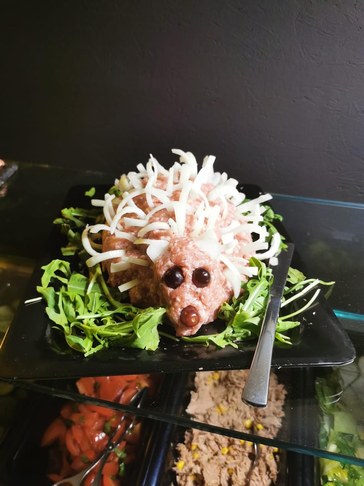Hackbraten-Igel mit Zwiebelstacheln, olivgrünen Augen und Rucola auf einem schwarzen Teller.