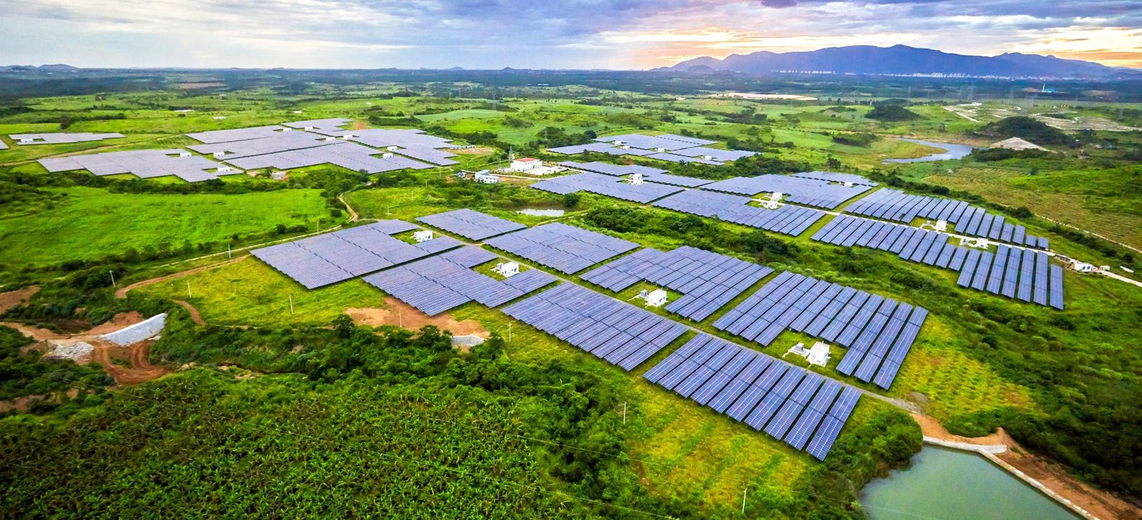 Vue aérienne d'une centrale solaire avec des rangées de panneaux bleus dans un paysage rural verdoyant au coucher du soleil.