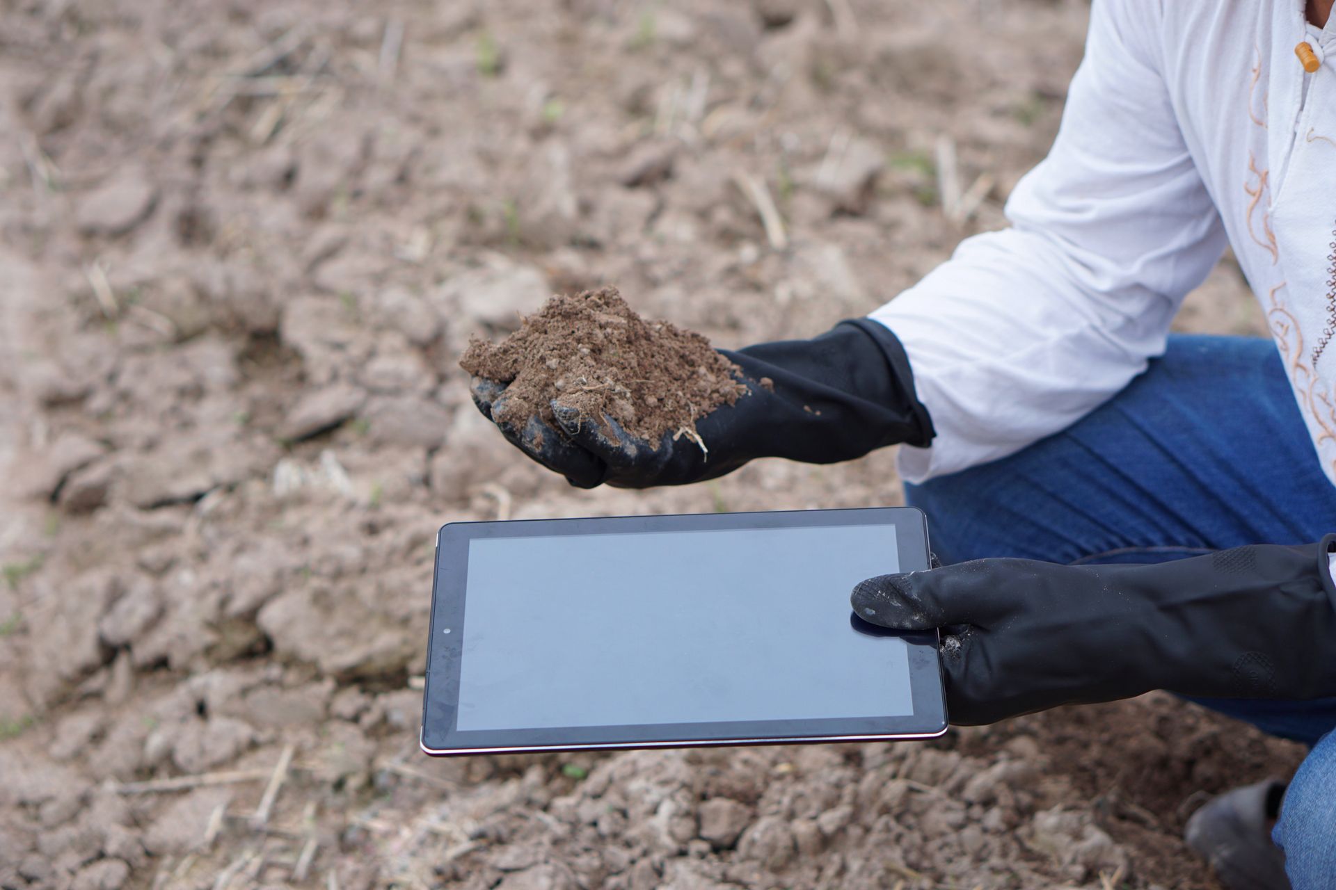 Des mains gantées tiennent une tablette et une pelletée de terre dans un champ.