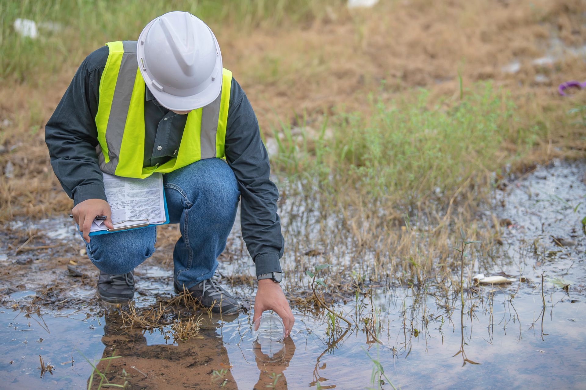Ouvrier portant un casque et un gilet de sécurité, agenouillé dans une zone humide boueuse, inspectant l'eau avec un outil.