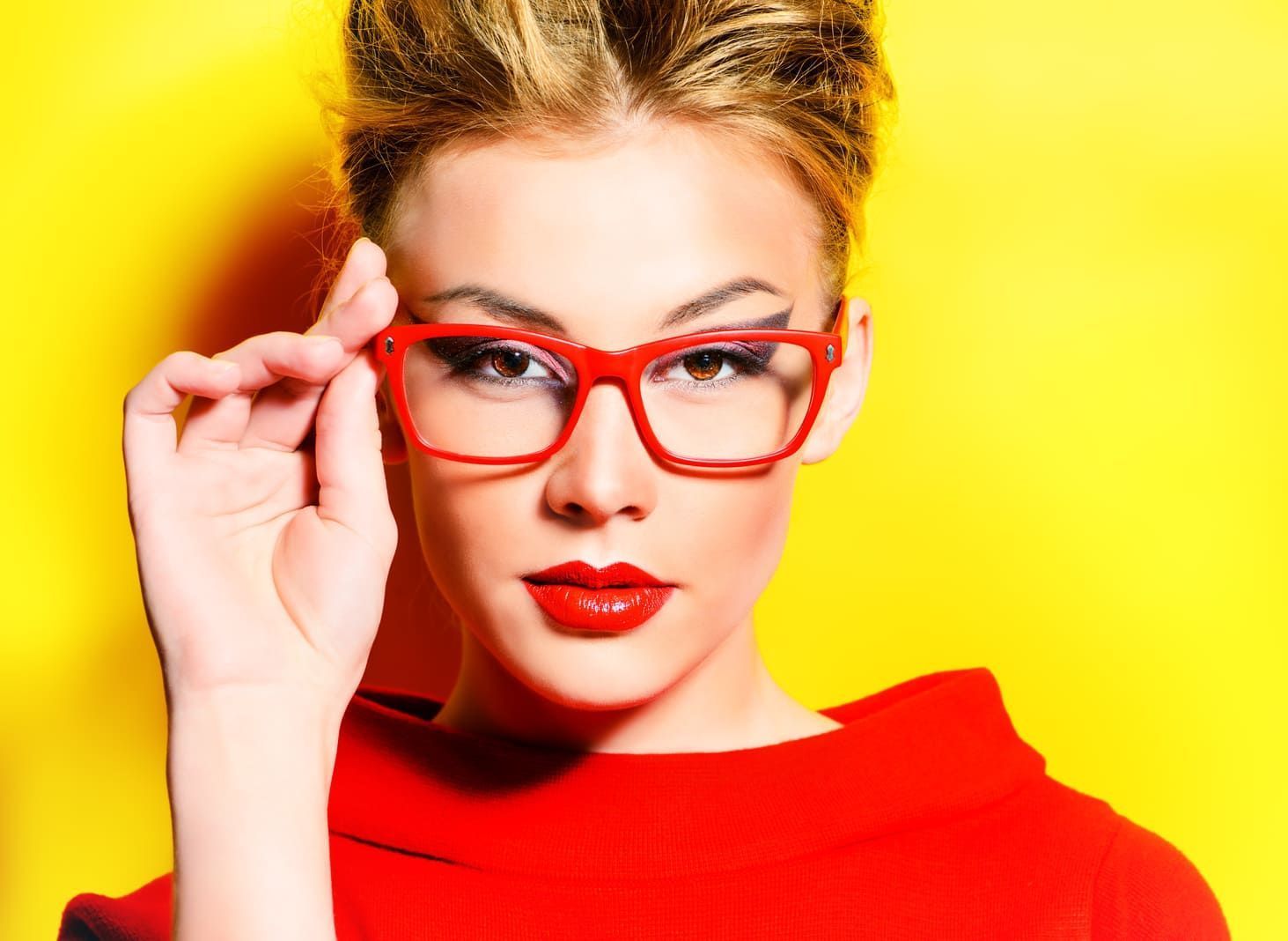 Una mujer que lleva gafas rojas y lápiz labial rojo sobre un fondo amarillo.