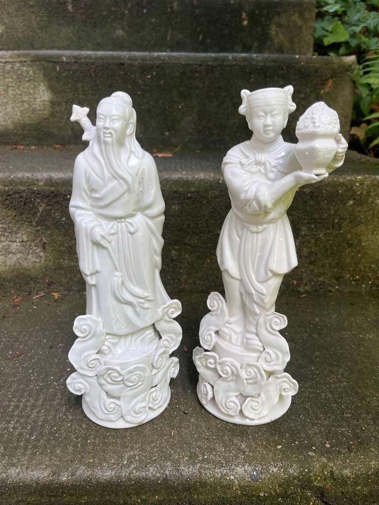 Deux statues en porcelaine blanche, l'une représentant un homme barbu avec un bâton, l'autre une femme tenant un bol, sur une base nuageuse.