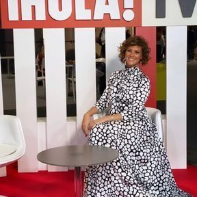 Una mujer con un vestido blanco y negro está sentada frente a un cartel que dice hola tv.