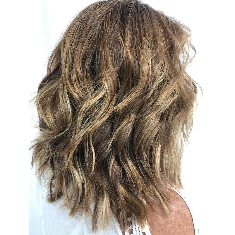 Una mujer con cabello castaño largo y ondulado lleva una camisa blanca.