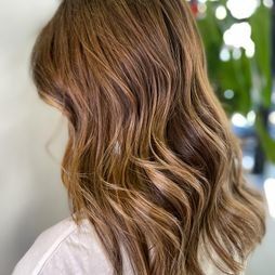 Una mujer con cabello castaño largo y ondulado lleva una camisa blanca.