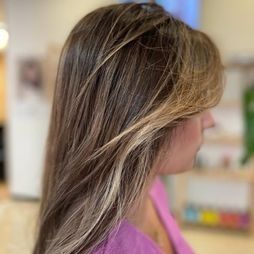 Una mujer con cabello largo castaño y reflejos rubios lleva una camisa rosa.