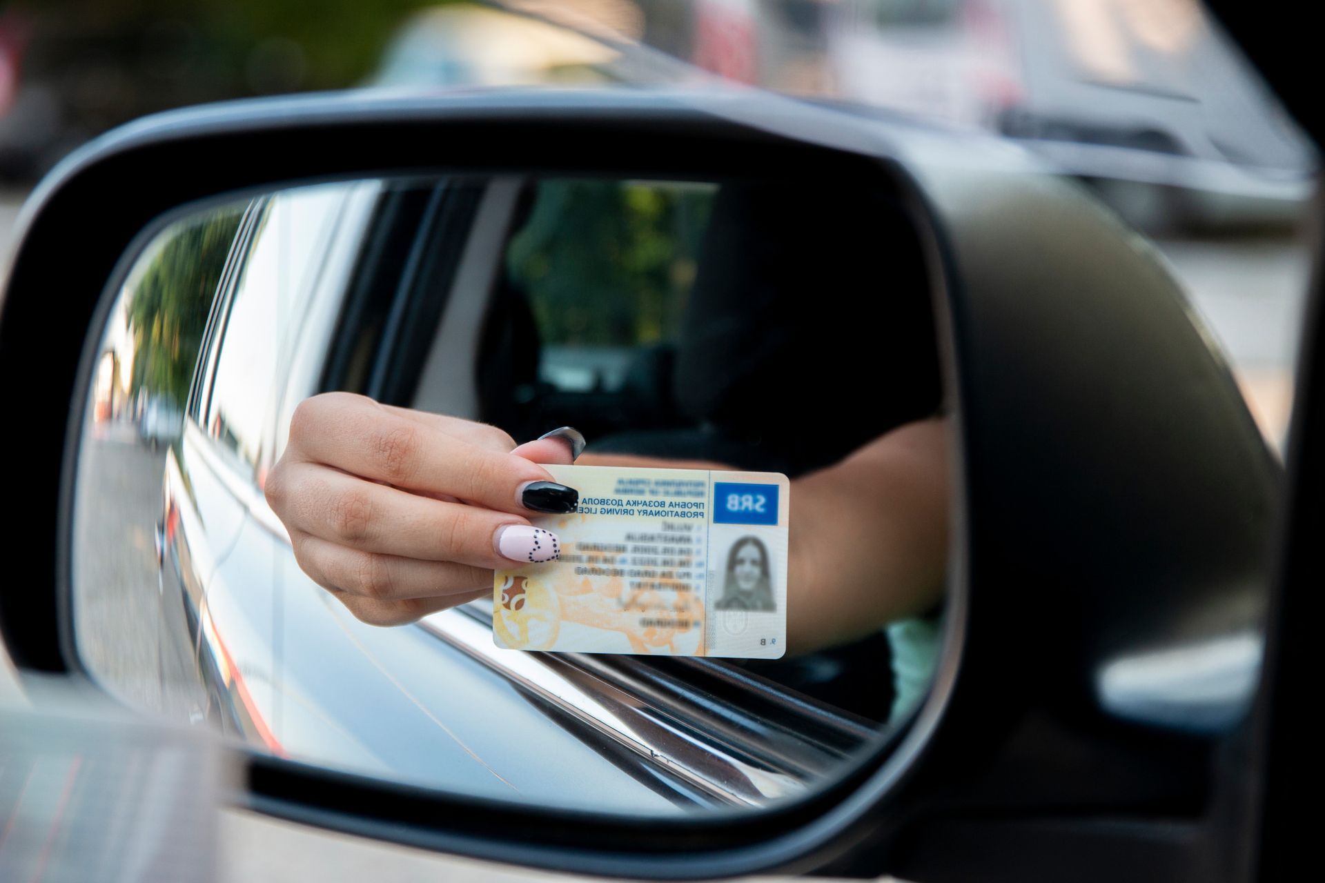 Una mujer sostiene una licencia de conducir frente al espejo retrovisor de un automóvil.