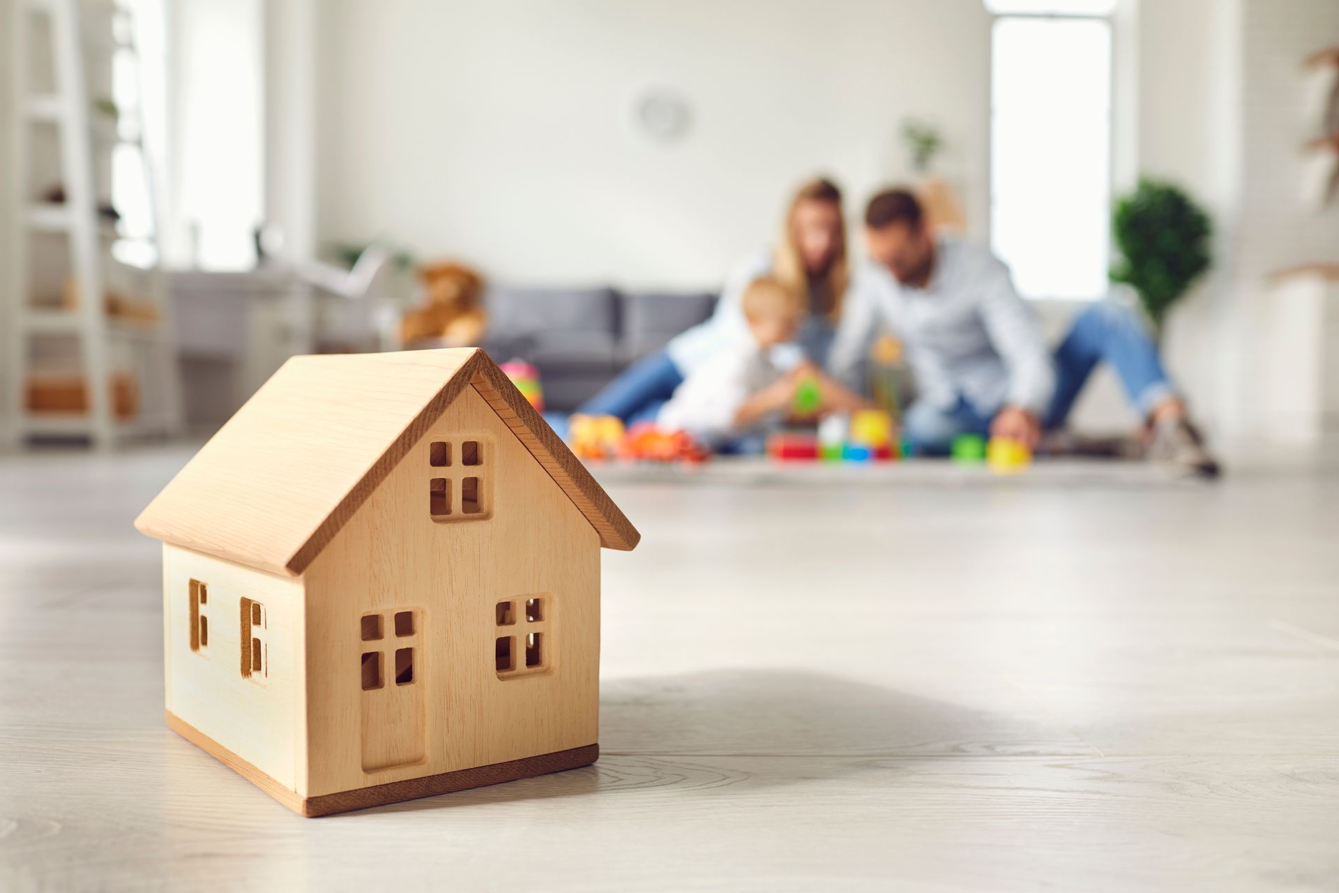 Au premier plan, une petite maison en bois ; à l'arrière-plan, des parents jouent avec leur enfant sur le sol du salon.