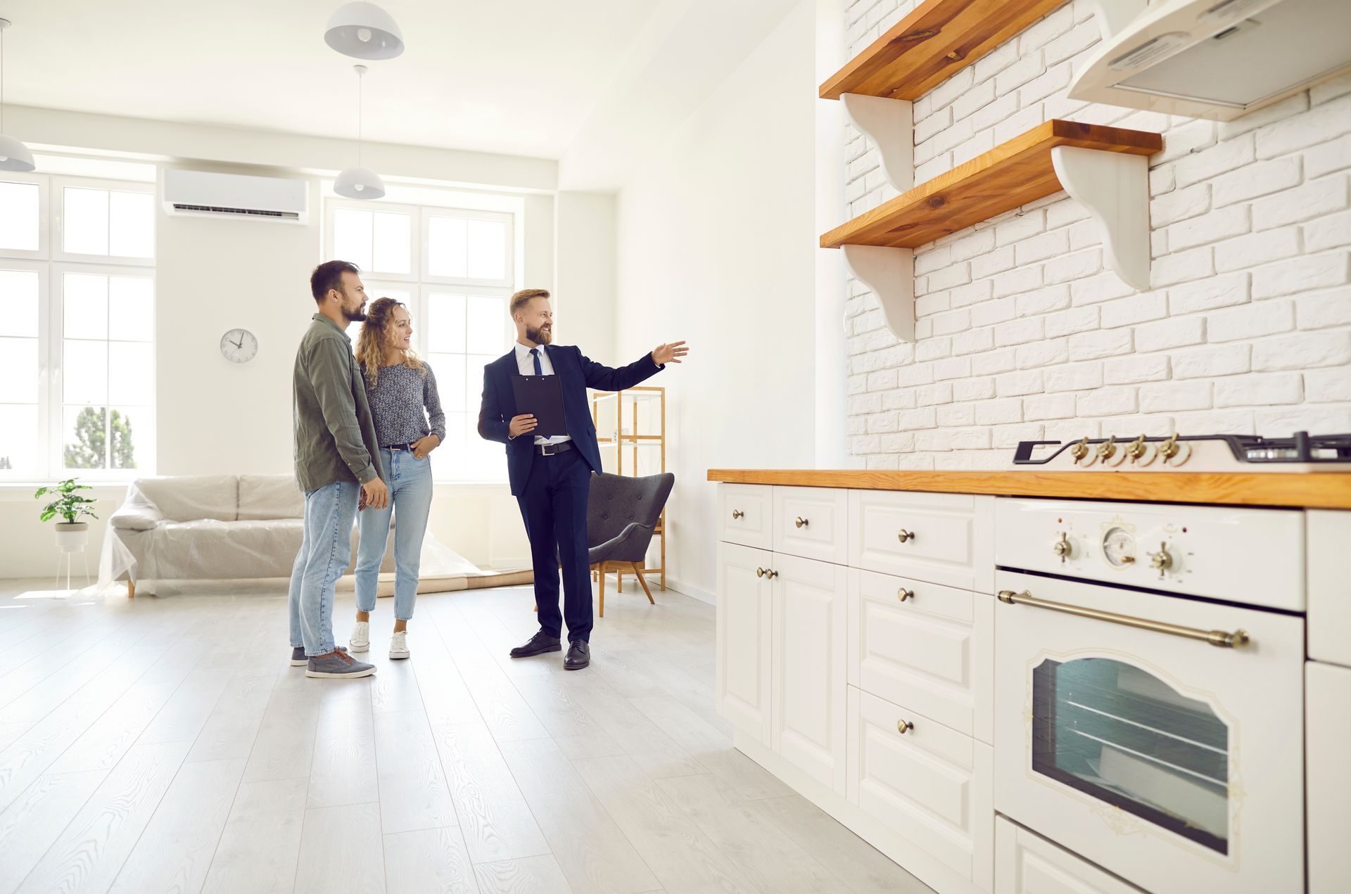 Deux personnes visitent une cuisine moderne et lumineuse tandis qu'un agent immobilier en présente les caractéristiques.