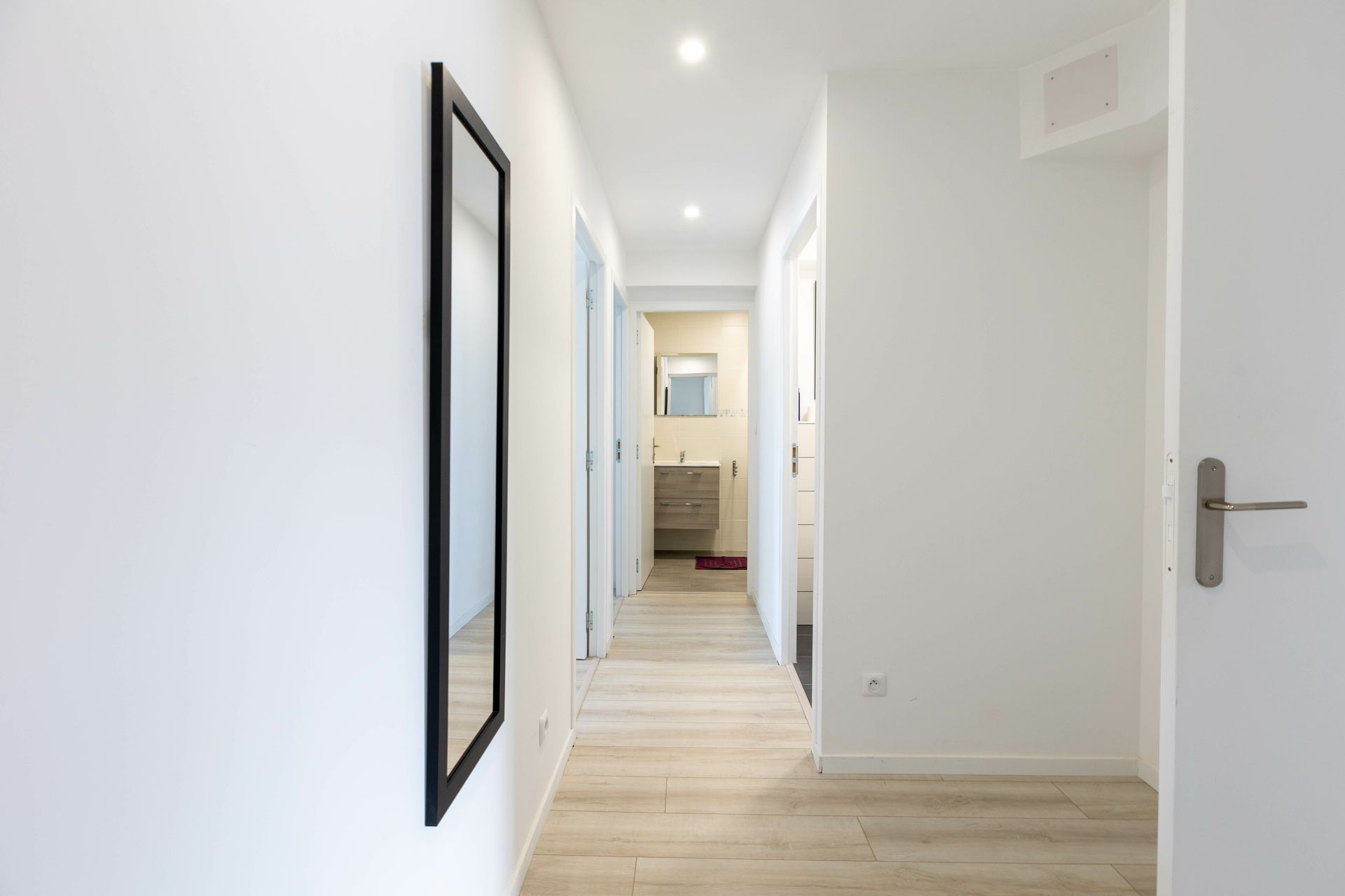 Couloir blanc lumineux avec parquet, spots encastrés, miroir et porte ouverte donnant sur une salle de bains.